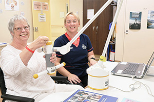 An older woman sits at a desk using a computer-based rehabilitation device. A female occupational therapist sits beside her