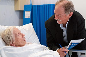 A man leans down to speak with an older woman laying in a hospital bed
