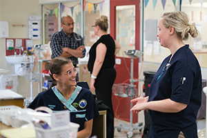 A woman wearing a Rockingham General Hospital uniform that reads 'Registered nurse' talks to another woman. Another man and woman stand talking in the background. 