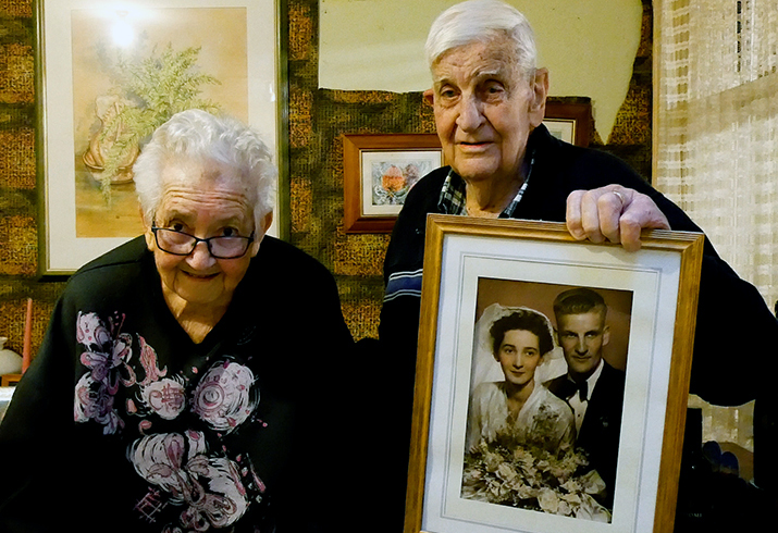 An older woman and man stand beside each other. The man holds a wedding photo of him and the woman.