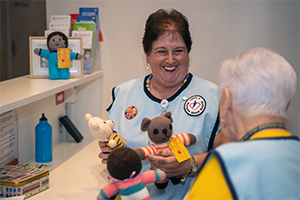 A woman wearing a tunic that has a badge reading Friends of Rockingham General Hospital  holds some knitted toys