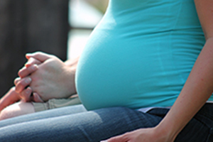 Close up photo of a pregnant woman's stomach. She is seated and has her left hand on the side of her left thigh. Another person is seated next to her with their hands clasped on their thighs.