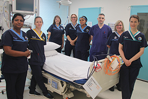 Eight people in nursing and medical scrubs stand in a treatment room beside a hospital bed