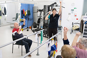 A female therapist demonstrates an arm exercise to three older people in a therapy gym.