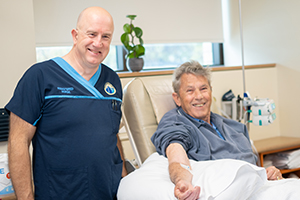 A male nurse stands beside an older man in a chair who is receiving chemotherapy treatment