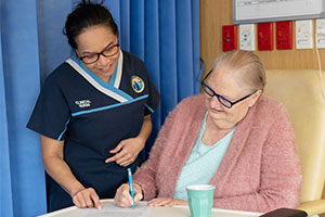 A female clinical nurse stands beside an older female patient who is signing a document