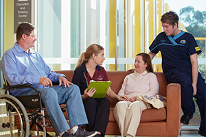 A male patient sites in a wheelchair. On a couch beside him sits a two health professionals and another woman