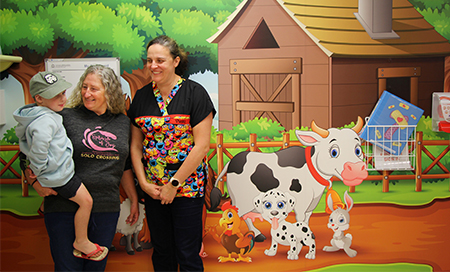 Grandmother holds grandson besides nurse in front of farm yard mural.