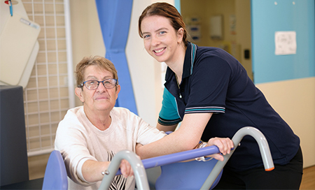 A young female physiotherapist supports and older woman to use a piece of therapy equipment.