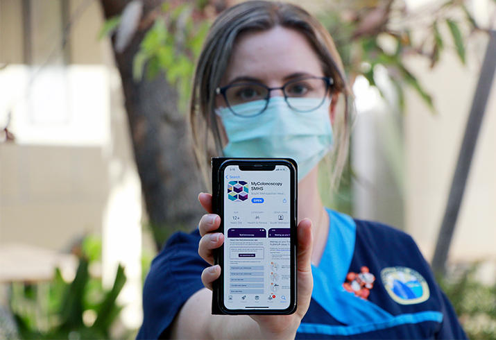 A female nurse wearing a surgical mask holds a smartphone displaying the MyColonscopy app on its screen