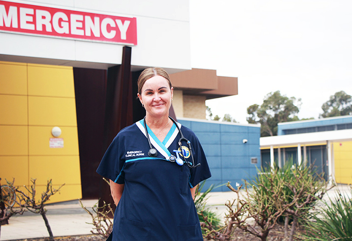 A woman in nursing scrubs stands outside a building. The word Emergency is on a sign across the top of the building.