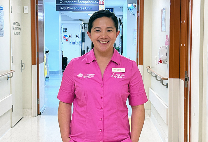 A registered nurse stands in a hospital corridor. She is wearing bright pink scrubs that feature the words McGrath Foundation