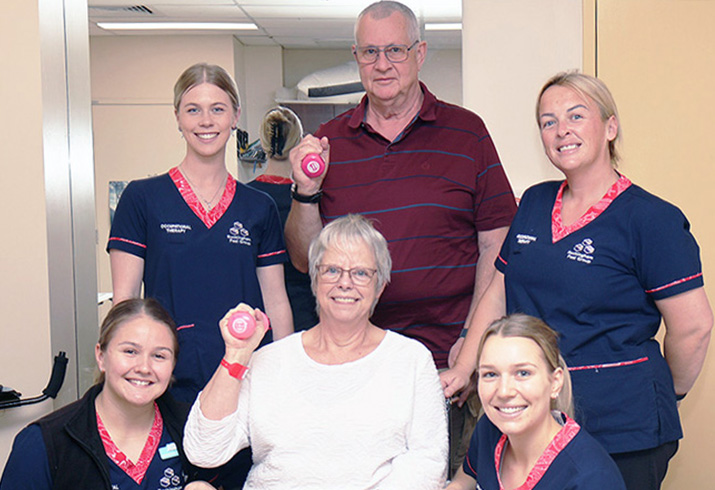An older woman sits holding a handweight in her right hand. Four female occupational therapists and an older gentleman stand around and behind her