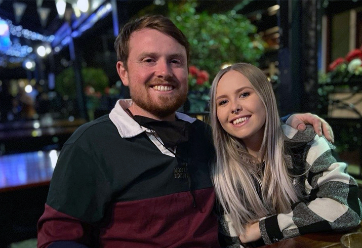 A young man sits with his arm around a young woman seated beside him