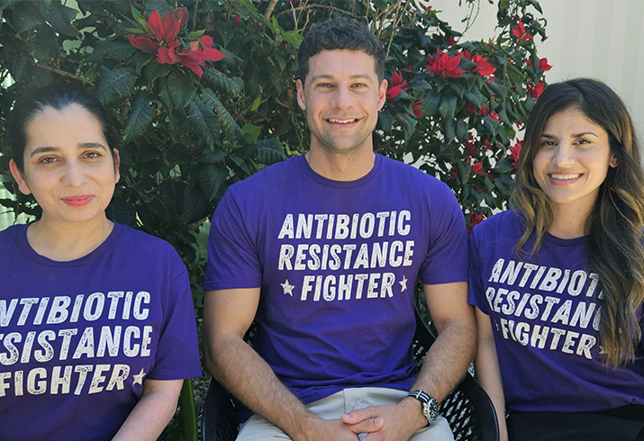 A man sits between two women in a garden. All three wear a shirt that reads 'Antibiotic resistance fighter'