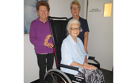 An elderly woman sits in a wheelchair. Behind her stands another woman and a female nurse.